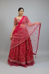 A woman modeling a traditional Indian red lehenga choli set with a sheer pink dupatta, photographed against a solid gray studio background.