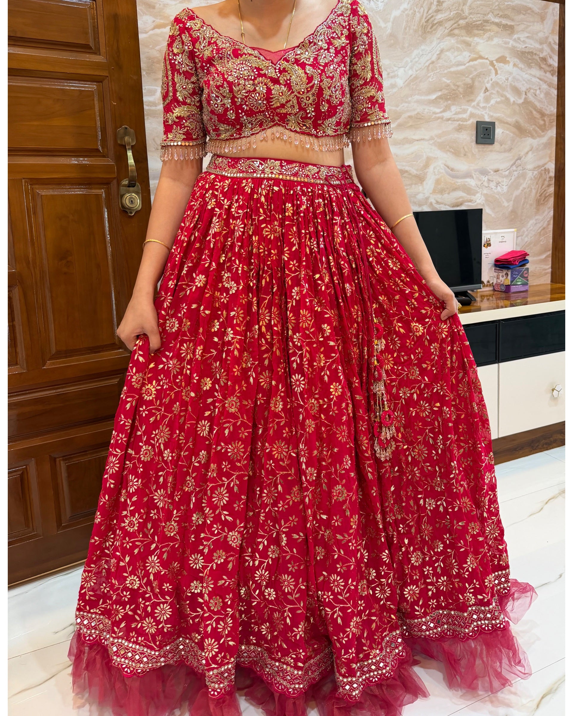 A red and gold embroidered traditional Indian lehenga choli set worn by a model in an indoor setting.