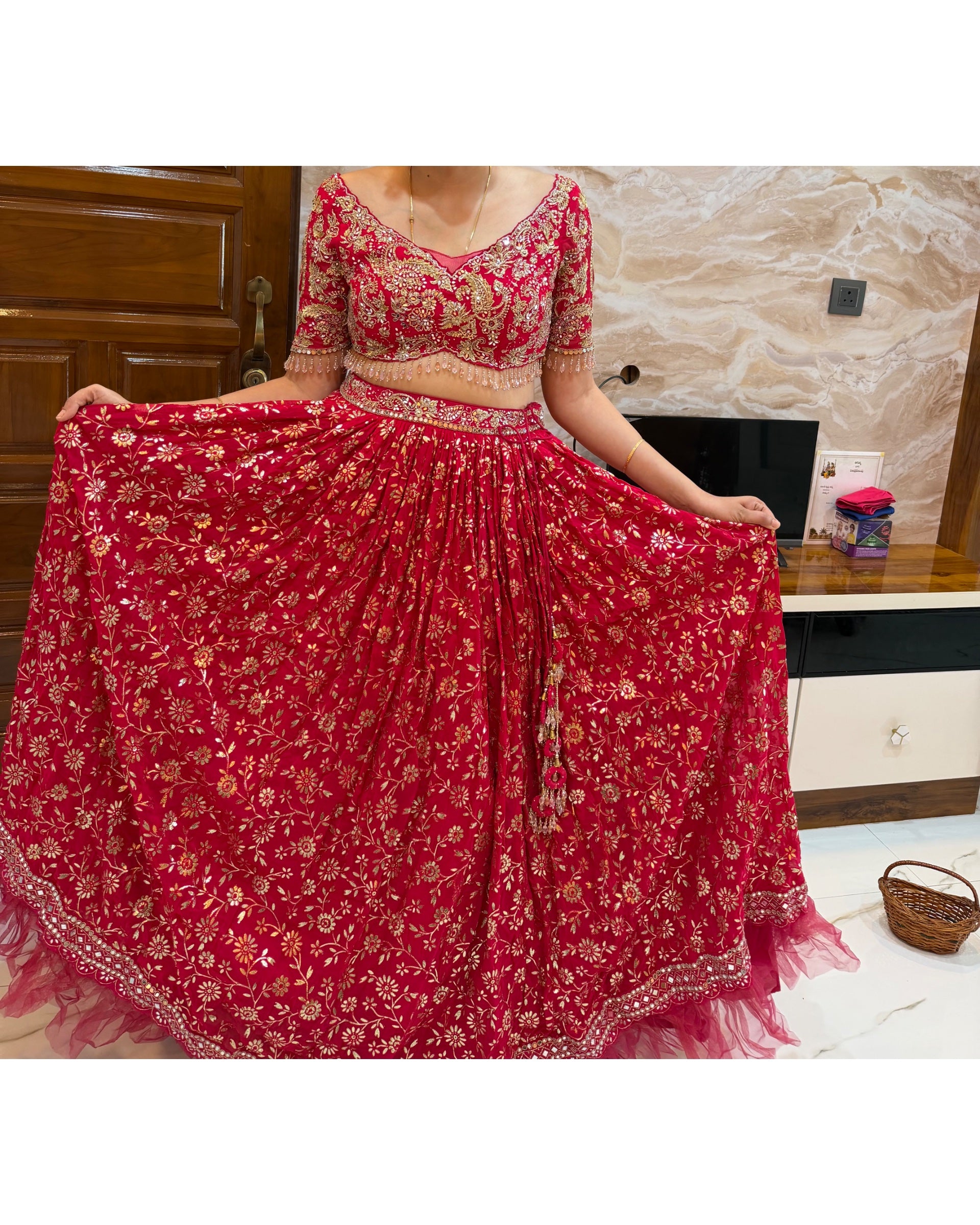 A red and gold floral embroidered lehenga choli set worn by a model, holding out the skirt to display its full volume in an indoor setting.