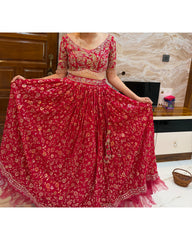 A red and gold floral embroidered lehenga choli set worn by a model, holding out the skirt to display its full volume in an indoor setting.