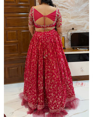 A woman viewed from the back wearing a red and gold embroidered traditional lehenga with a flared skirt and embellished blouse, standing indoors in front of a wooden door and TV stand.