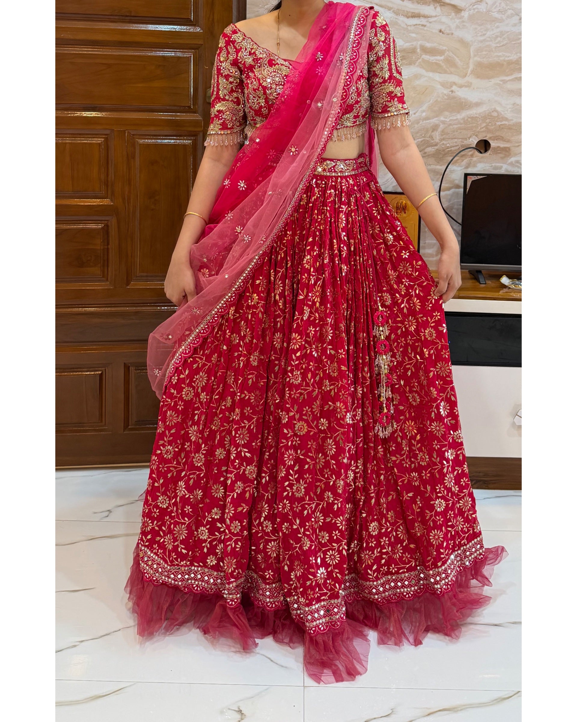 A woman wearing a bright pink and red floral lehenga choli set with a matching dupatta, standing indoors in front of a wooden door and marble floor.