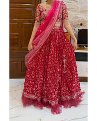 A woman wearing a bright pink and red floral lehenga choli set with a matching dupatta, standing indoors in front of a wooden door and marble floor.