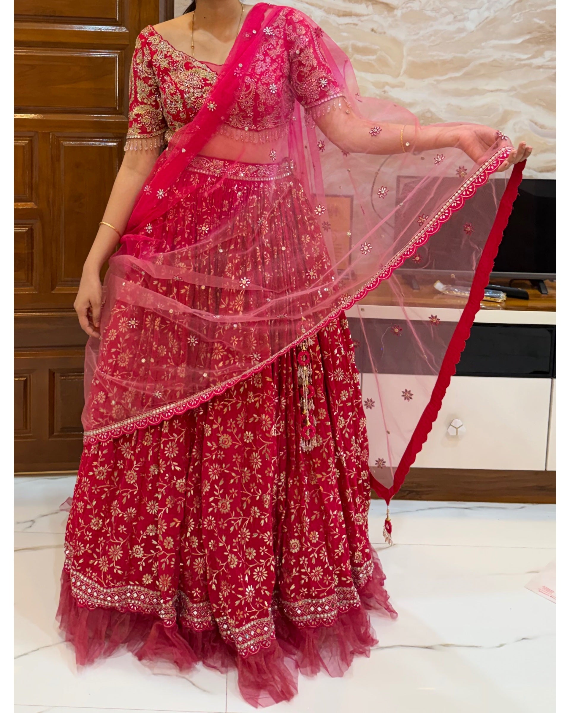 A woman wearing a bright pink and red floral lehenga choli set with a sheer dupatta, standing indoors in front of a wooden door and marble wall.