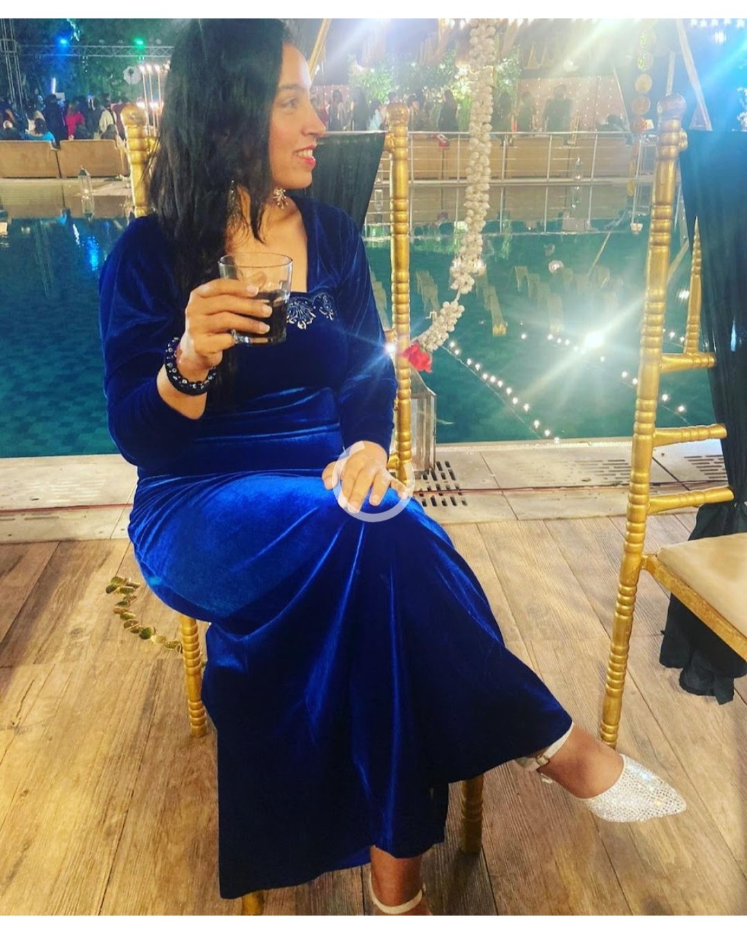 Woman in a blue dress sitting on a chair holding a glass, with a festive background.