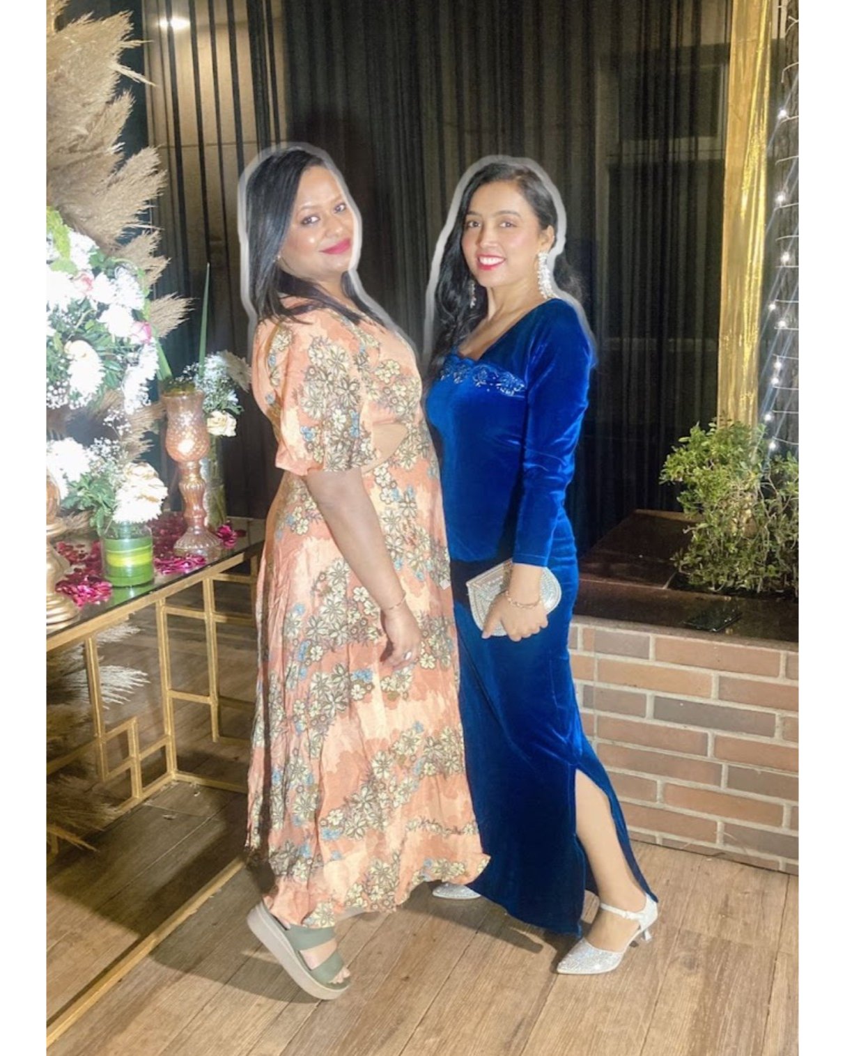Two women posing together in a decorated indoor setting with plants and decor.