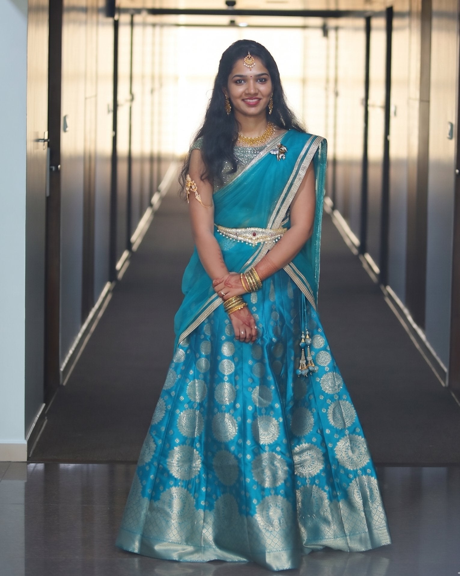 Woman in a blue half saree with silver patterns standing in a hallway.